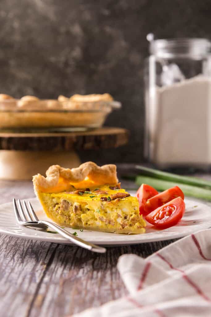 side shot of a slice of quiche. Small chunks of ham can be seen in the custard. Juicy red tomatoes have been cut and are served alongside the quiche.