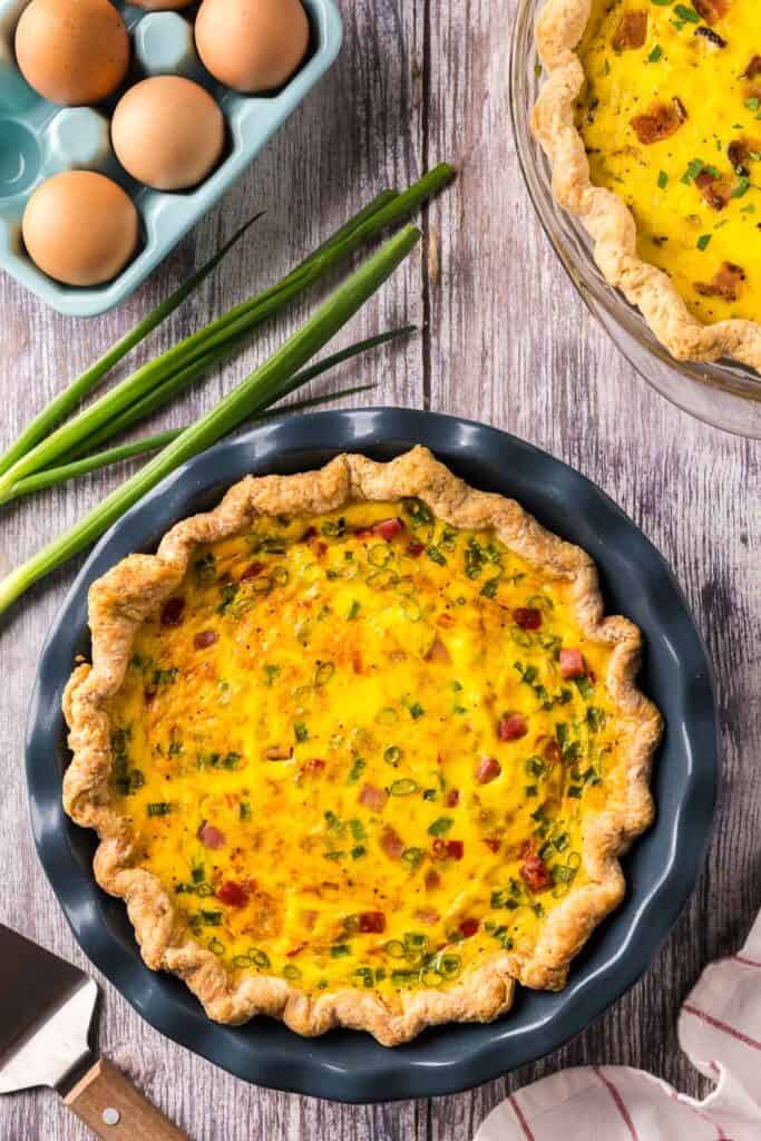 An overhead shot of a ham and cheese quiche in a blue pie dish. A blue container of eggs and a pie server are in the background.