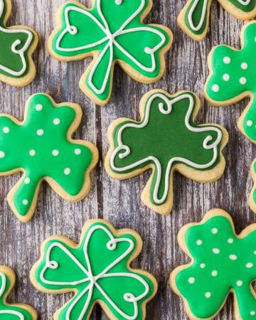 Overhead horizontal close up shot of shamrock-shaped sugar cookies laid out on a wooden table. There are three different decorated variations of the same shape cookie.