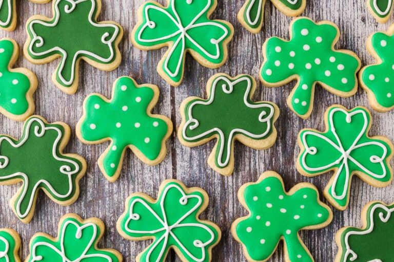 Overhead horizontal close up shot of shamrock-shaped sugar cookies laid out on a wooden table. There are three different decorated variations of the same shape cookie.