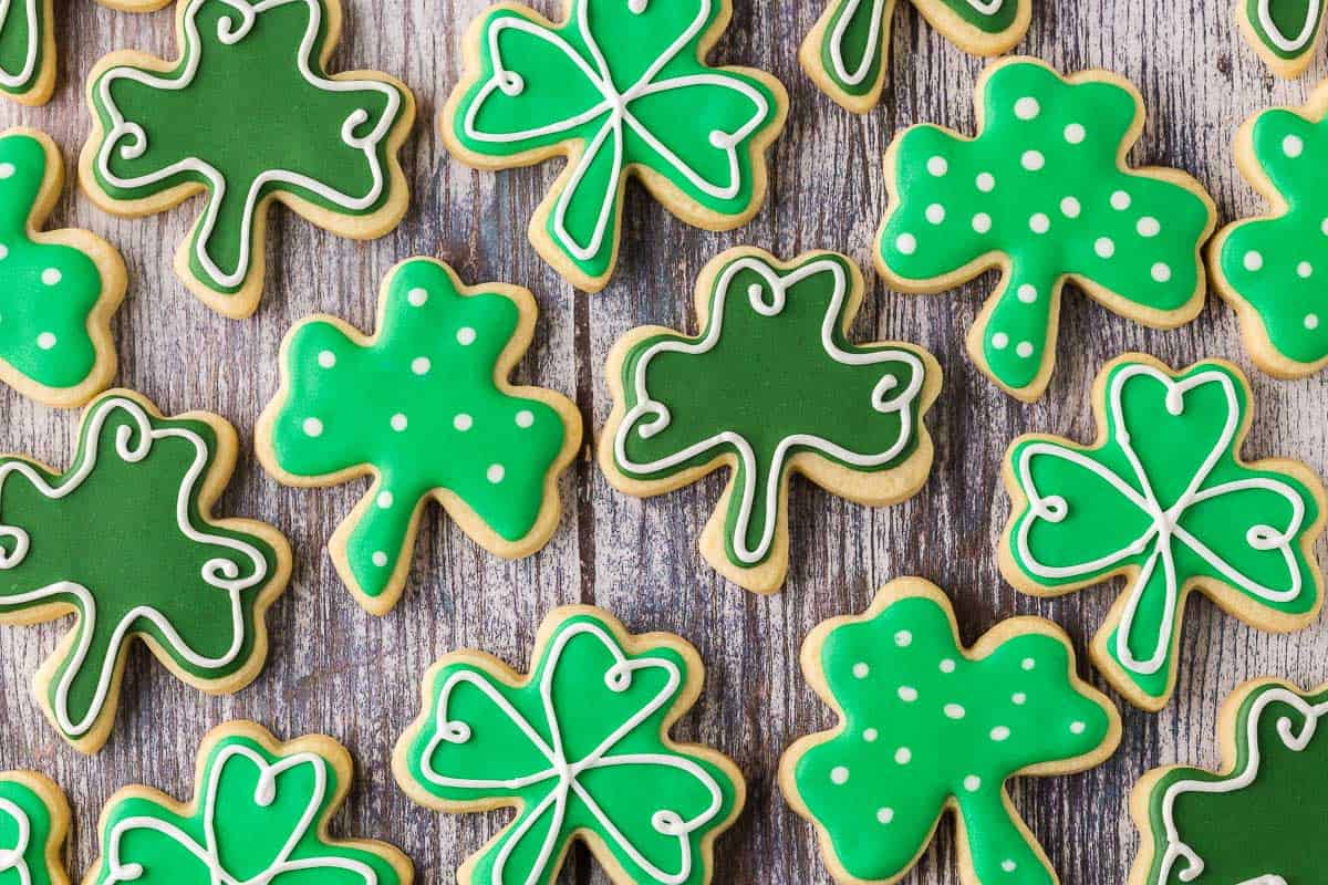 Overhead horizontal close up shot of shamrock-shaped sugar cookies laid out on a wooden table. There are three different decorated variations of the same shape cookie.
