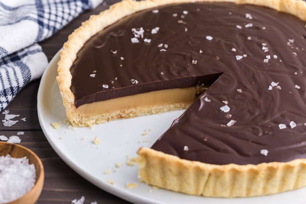 low angle shot of a chocolate caramel tart with a slice removed.  The removal of the slice exposes the tarts three layers, the tart crust, followed by a golden caramel layer, and topped with a dark chocolate ganache.