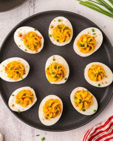 Black plate with devilled eggs on it. The devilled eggs all point outwards along the edge of the plate except for one egg in the middle. All have had the devilled egg filling piped into the middle of the egg and topped with paprika and finely chopped chives.