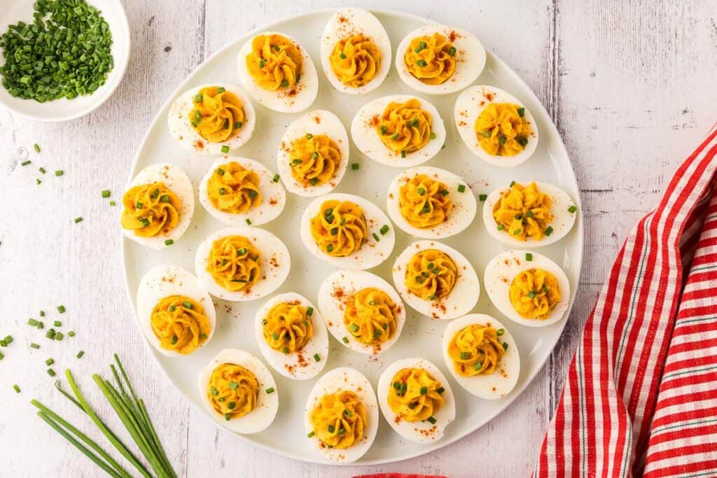 overhead shot of a large white platter filled with devilled eggs.  Each devilled egg has had the filling piped into the center and garnished with paprika and fresh cut chives.