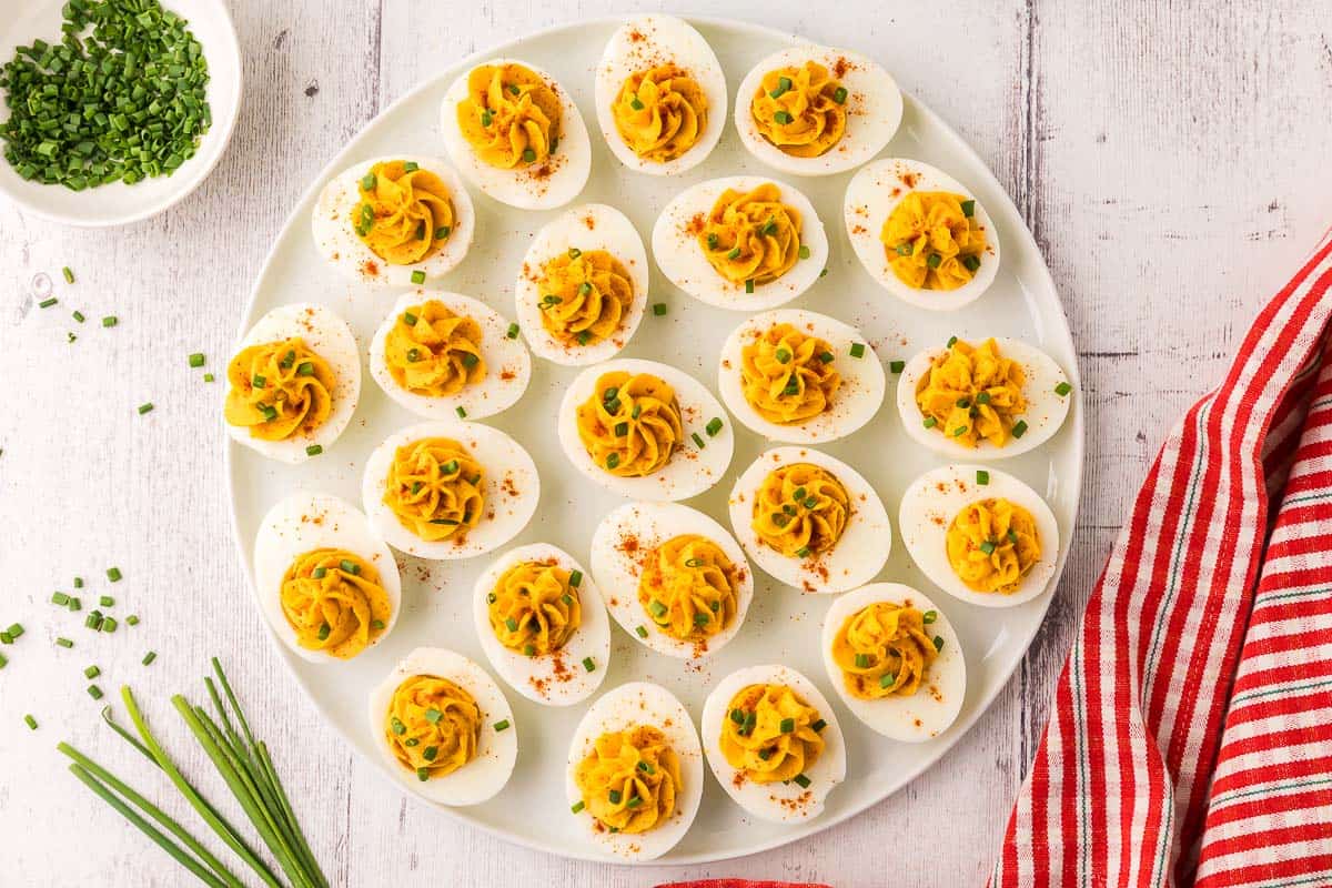 overhead shot of a large white platter filled with devilled eggs. Each devilled egg has had the filling piped into the center and garnished with paprika and fresh cut chives. These go fast at a brunch.