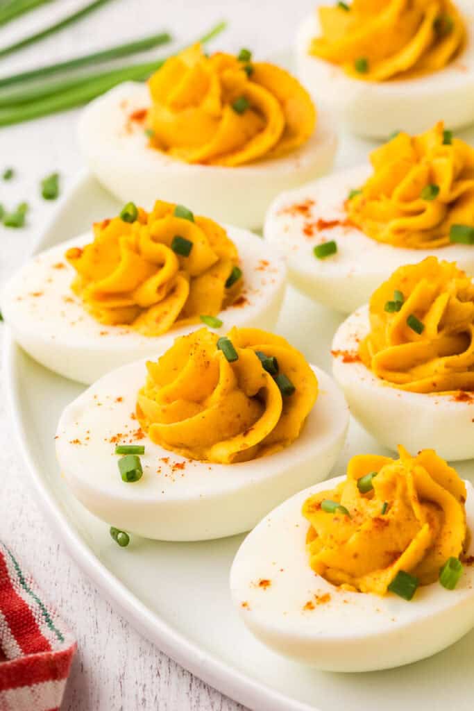 A side shot of a white serving plate with devilled eggs. Each egg has been nicely piped with the filling and sprinkled with paprika and chopped chives as garnish