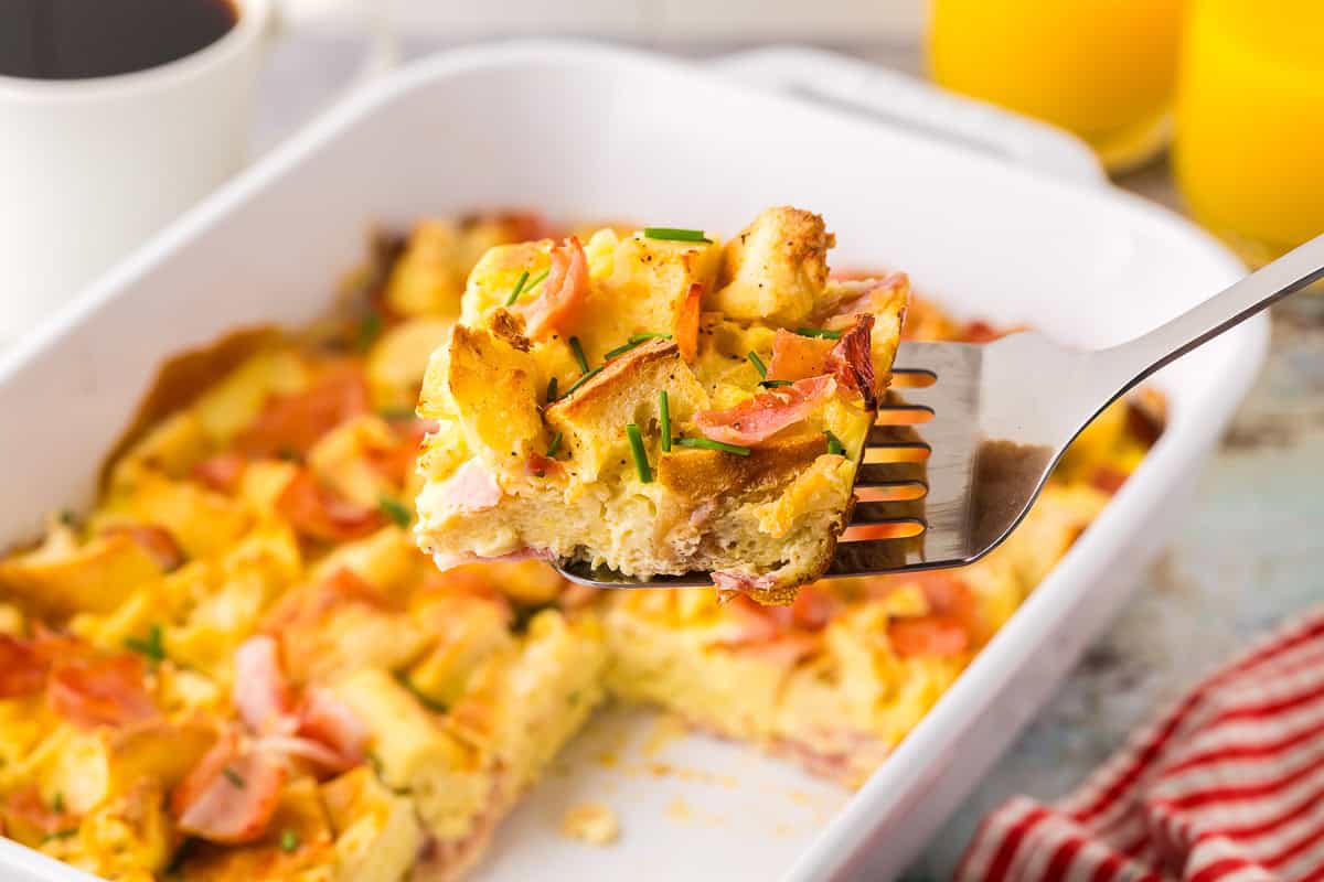 horizontal side shot looking at a slice of eggs benedict casserole being removed from the casserole dish with a metal spatula. The square slice is made with chunks of English muffin, and Canadian ham, held together with custardy egg. Chives have been sprinkled on top. A great recipe if you are hosting brunch.