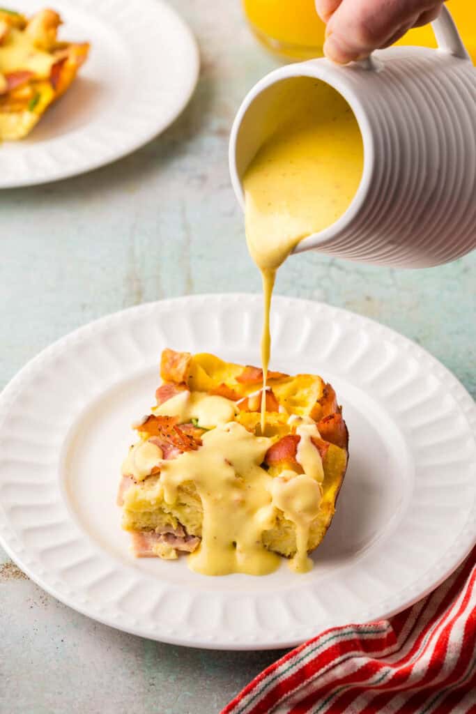 Side shot of a small white jug pouring hollandaise sauce over a square slice of eggs benedict casserole.