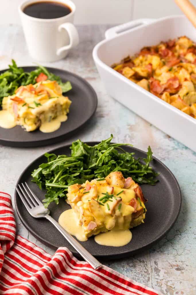 45 degree angle shot of a dark grey plate with a square slice of eggs benedict casserole drizzled with hollandaise sauce.  There is a small bunch of arugula tossed in a vinaigrette on the plate as well.