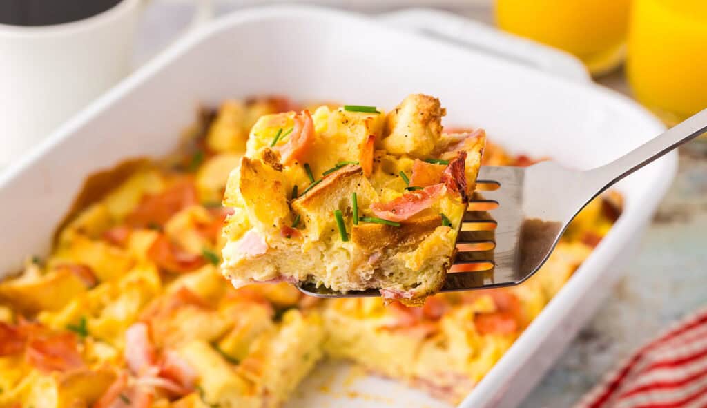 horizontal side shot looking at a slice of eggs benedict casserole being removed from the casserole dish with a metal spatula. The square slice is made with chunks of English muffin, and Canadian ham, held together with custardy egg. Chives have been sprinkled on top.