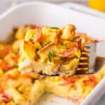 horizontal side shot looking at a slice of eggs benedict casserole being removed from the casserole dish with a metal spatula. The square slice is made with chunks of English muffin, and Canadian ham, held together with custardy egg. Chives have been sprinkled on top.