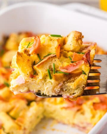 horizontal side shot looking at a slice of eggs benedict casserole being removed from the casserole dish with a metal spatula. The square slice is made with chunks of English muffin, and Canadian ham, held together with custardy egg. Chives have been sprinkled on top.