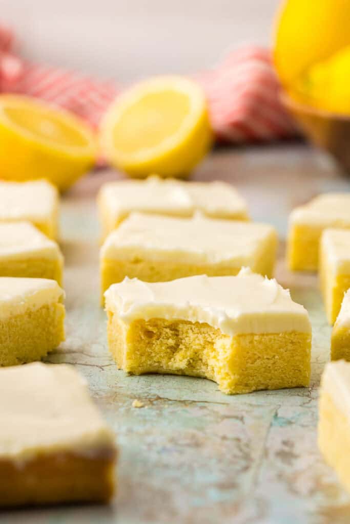 Side shot looking at a lemon cookie bar topped with cream cheese frosting with a bite out of it. It is sitting on the counter surrounded by other lemon cookie bars, and in the background are more lemons and a red linen.