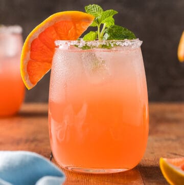 Close up side shot of a paloma mocktail in a non-stemmed wine glass. It is a light pink in color and garnished with a grapefruit wedge on the edge of the glass and a sprig of mint. The glass is filled with ice and has a salted rim. There are two more paloma mocktails in the background.