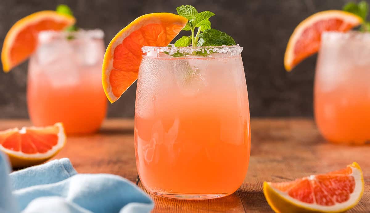 Close up side shot of a paloma mocktail in a non-stemmed wine glass. It is a light pink in color and garnished with a grapefruit wedge on the edge of the glass and a sprig of mint. The glass is filled with ice and has a salted rim. There are two more paloma mocktails in the background.