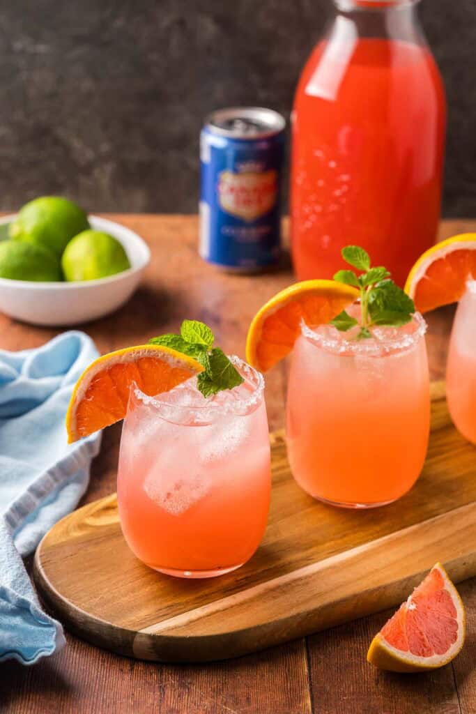 45 degree angle shot of paloma mocktails full of ice and garnished with grapefruit wedges and sprigs of mint.  Three of these pink drinks sit on a thin wood serving board at an angle.  In the background is a bowl of limes, jug of grapefruit juice, and a can of club soda.