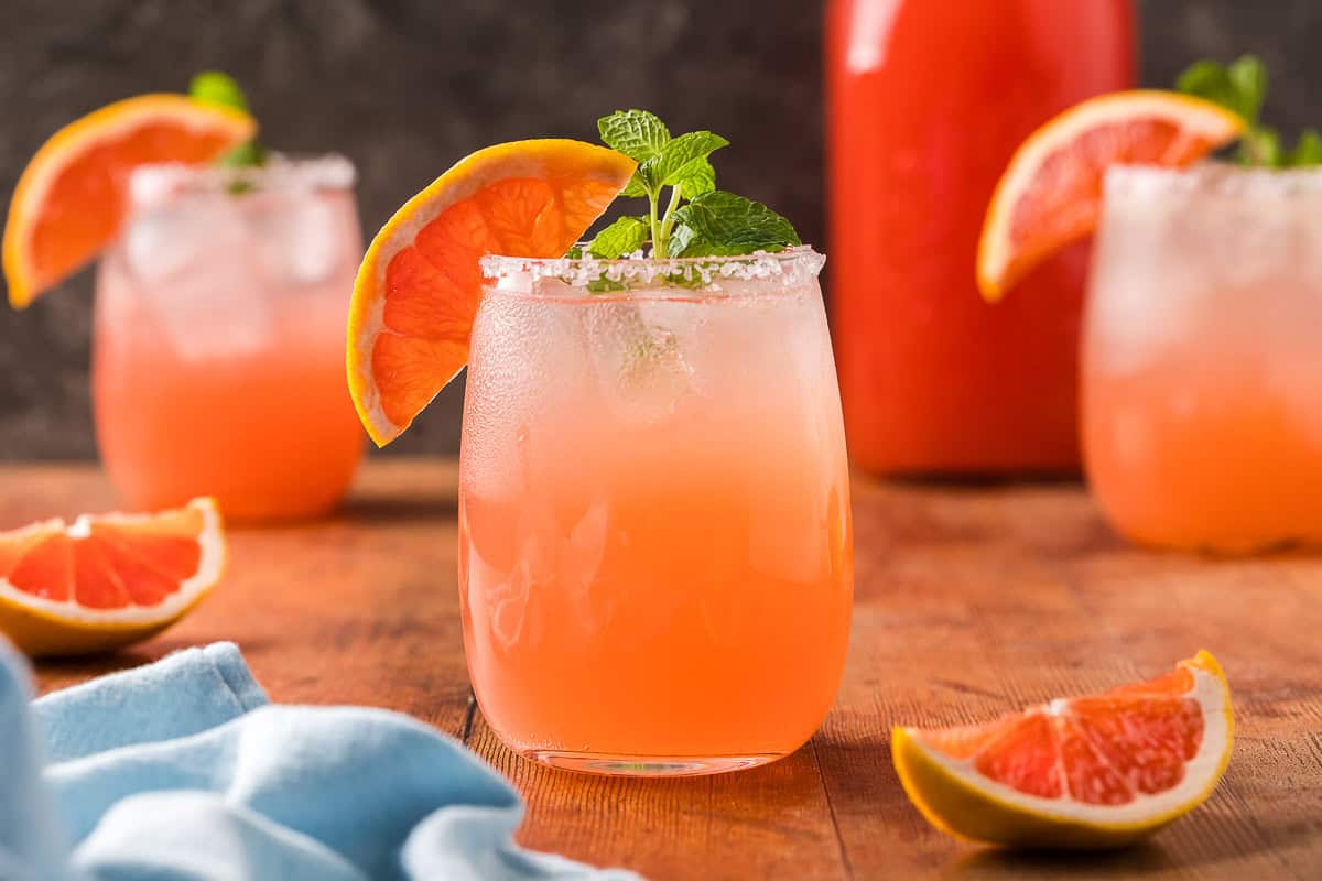 Close up side shot of a paloma mocktail in a non-stemmed wine glass. It is a light pink in color and garnished with a grapefruit wedge on the edge of the glass and a sprig of mint. The glass is filled with ice and has a salted rim. There are two more paloma mocktails in the background along with a carafe of grapefruit juice.