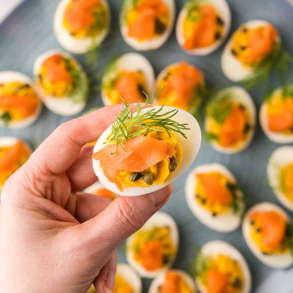 Overhead shot of a smoked salmon devilled egg being lifted up toward the camera. This view shows off the sprig of fresh dill, slice of smoked salmon, and capers, a top of the creamy piped filling.
