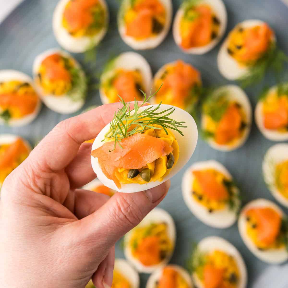 Overhead shot of a smoked salmon devilled egg being lifted up toward the camera. This view shows off the sprig of fresh dill, slice of smoked salmon, and capers, a top of the creamy piped filling.