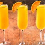 Horizontal side shot of three champagne flutes side by side filled with non-alcoholic orange mimosa. Half an orange wheel is on the edge of each glass.