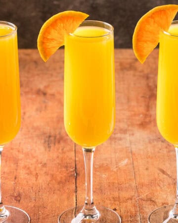 Horizontal side shot of three champagne flutes side by side filled with non-alcoholic orange mimosa. Half an orange wheel is on the edge of each glass.