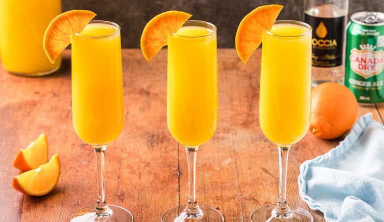 Horizontal side shot of three champagne flutes side by side filled with non-alcoholic orange mimosa. Half an orange wheel is on the edge of each glass.