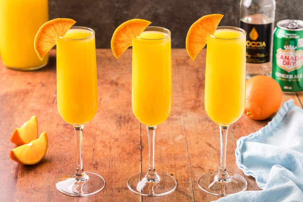 Horizontal side shot of three champagne flutes side by side filled with non-alcoholic orange mimosa. Half an orange wheel is on the edge of each glass.