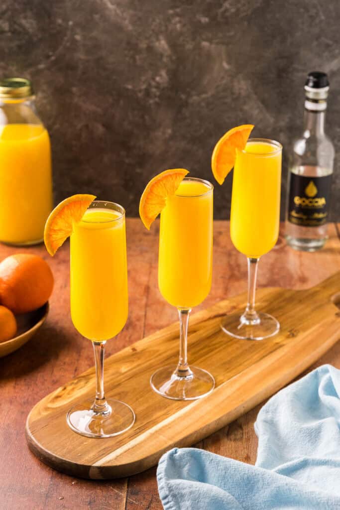 Side shot of three orange non-alcoholic mimosas on a narrow wooden serving platter.  Each is garnished with an orange wheel on the rim of the glass.