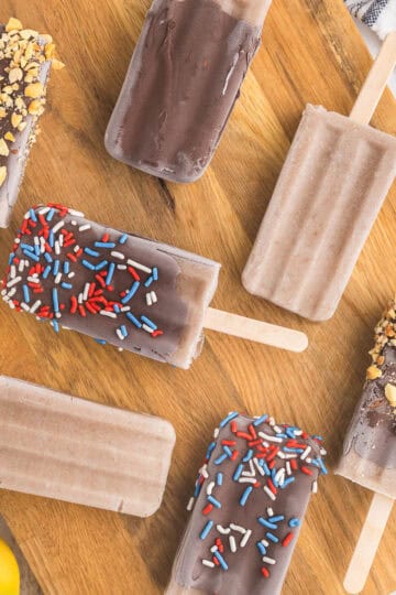seven banana popsicles laying askew on a wooden cutting board. Some are dipped in chocolate with crushed peanuts, others dipped in chocolate with red white and blue sprinkles and a few are plain.