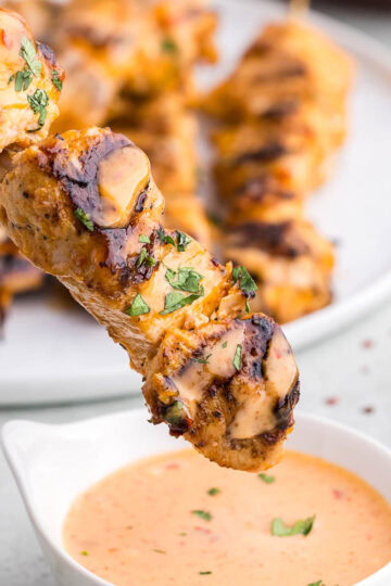 close up side shot of a Bang Bang Chicken Skewer with black grill marks and garnished with fresh chopped parsley. The skewer is just about to be dipped into a small bowl of bang bang sauce. In the background is a plate full of more chicken skewers.