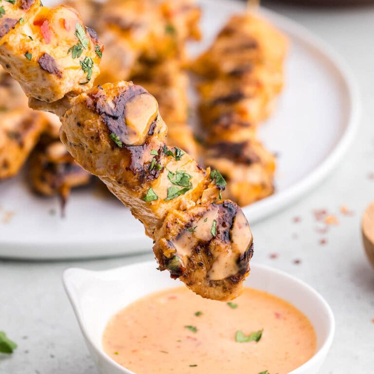 close up side shot of a Bang Bang Chicken Skewer with black grill marks and garnished with fresh chopped parsley. The skewer is just about to be dipped into a small bowl of bang bang sauce. In the background is a plate full of more chicken skewers.