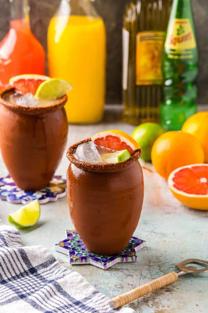Low angle shot of a cantarito cocktail served in the traditional clay cup rimmed with tajin. In the background are fresh grapefruit and lime as well as bottles of juice and tequila.