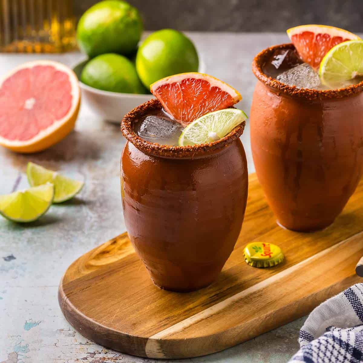 side shot of two Cantaritos cocktails in the traditional clay cantarito cup. They are rimmed with Tajin and garnished with grapefruit and lime wheels.