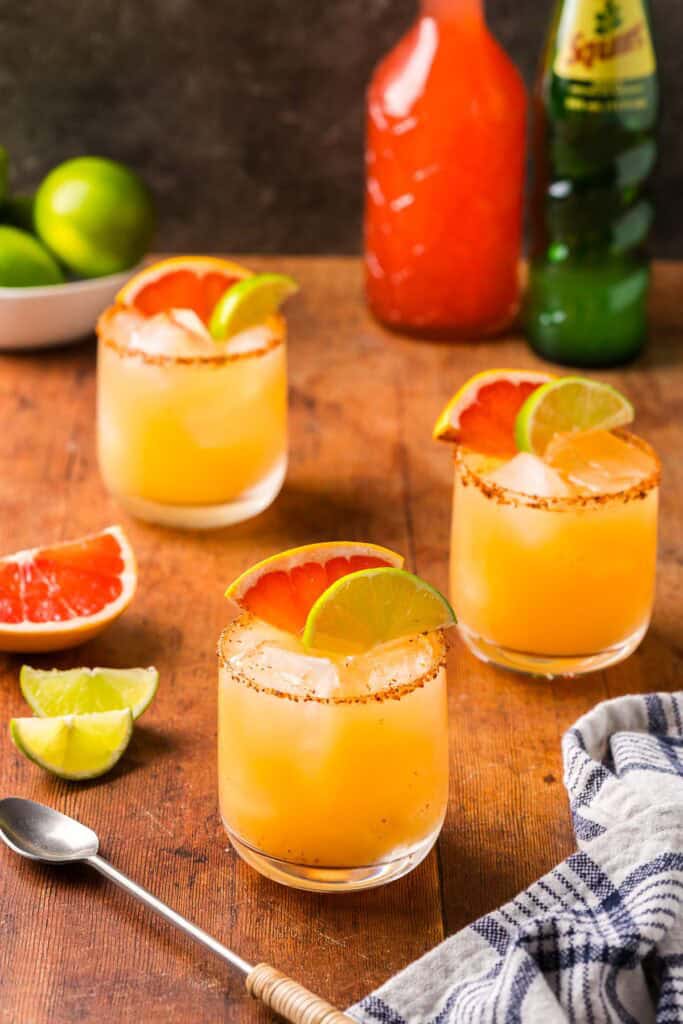 Low angle shot of three cantarito cocktails being served in lowball glasses. Each one is rimmed with Tajin and garnished with a portion of a grapefruit and lime wheel.