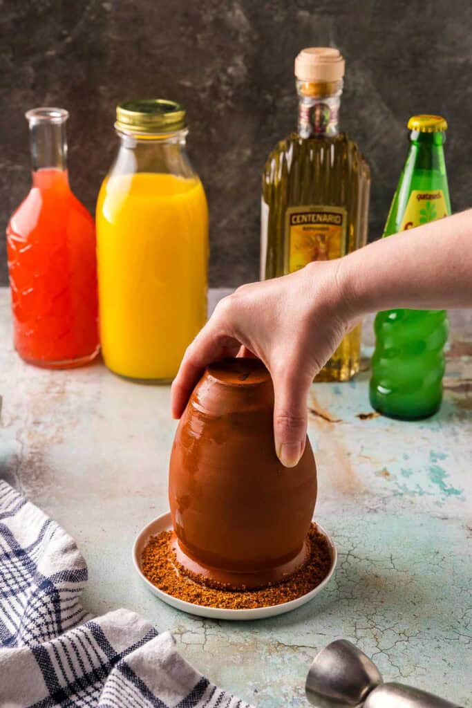 Side shot of a saturated cantarito clay cup being inverted onto a plate of Tajin in order to rim the cup.