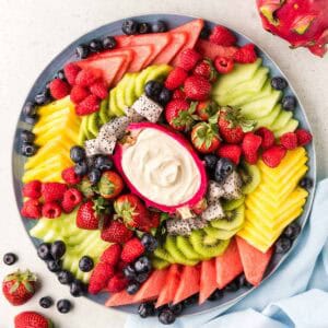 Overhead shot of a fruit platter. In the center is a hollowed out dragon fruit half filled with a creamy fruit dip, and around the the dip are small wedges of watermelon, pineapple, honey dew, kiwi rounds, blueberries, raspberries, and strawberries