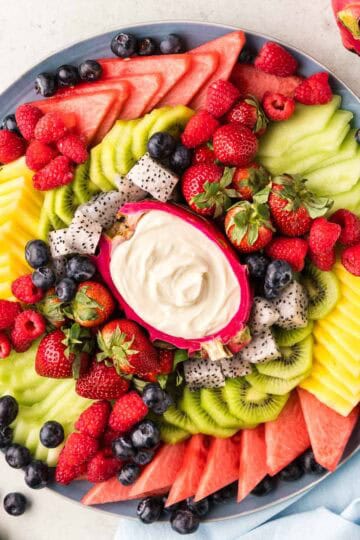 Overhead shot of a fruit platter. In the center is a hollowed out dragon fruit half filled with a creamy fruit dip, and around the the dip are small wedges of watermelon, pineapple, honey dew, kiwi rounds, blueberries, raspberries, and strawberries
