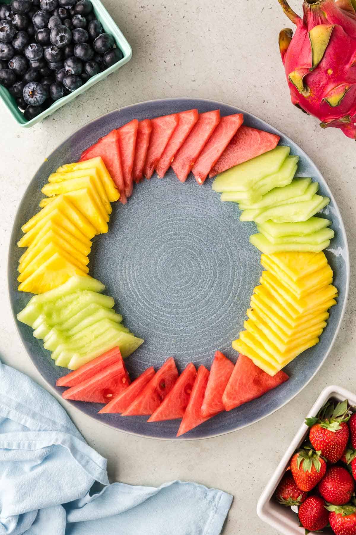 Overhead shot of a large blue circular serving plate. The large pieces of watermelon, honeydew, and pineapple are added in a ring around the outside edge of the plate.