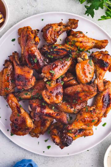 A white plate filled with hot honey chicken wings. The wings are a glossy red-brown and are garnished with chopped parsley. A little bowl of ranch dressing is available for dipping.