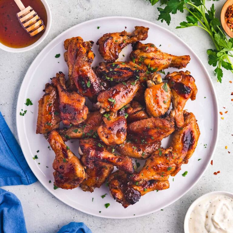 A white plate filled with hot honey chicken wings. The wings are a glossy red-brown and are garnished with chopped parsley. A little bowl of ranch dressing is available for dipping.