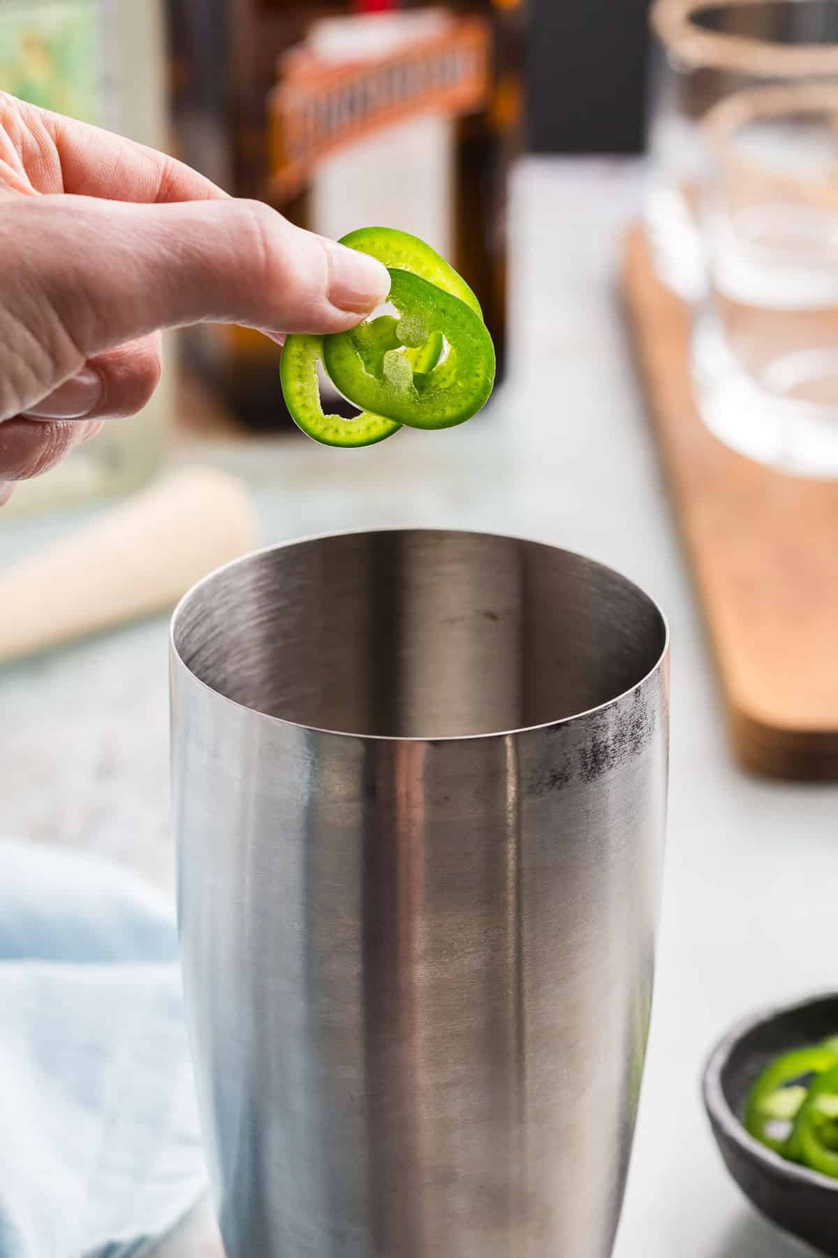 side shot of a stainless steel cocktail shaker having three jalapeno rounds being added to it.
