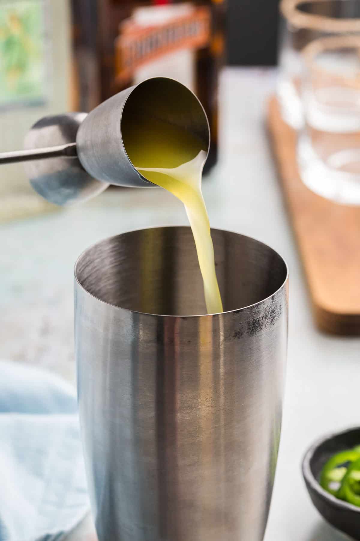 Side shot of a jigger pouring pineapple juice into a stainless steel cocktail shaker.