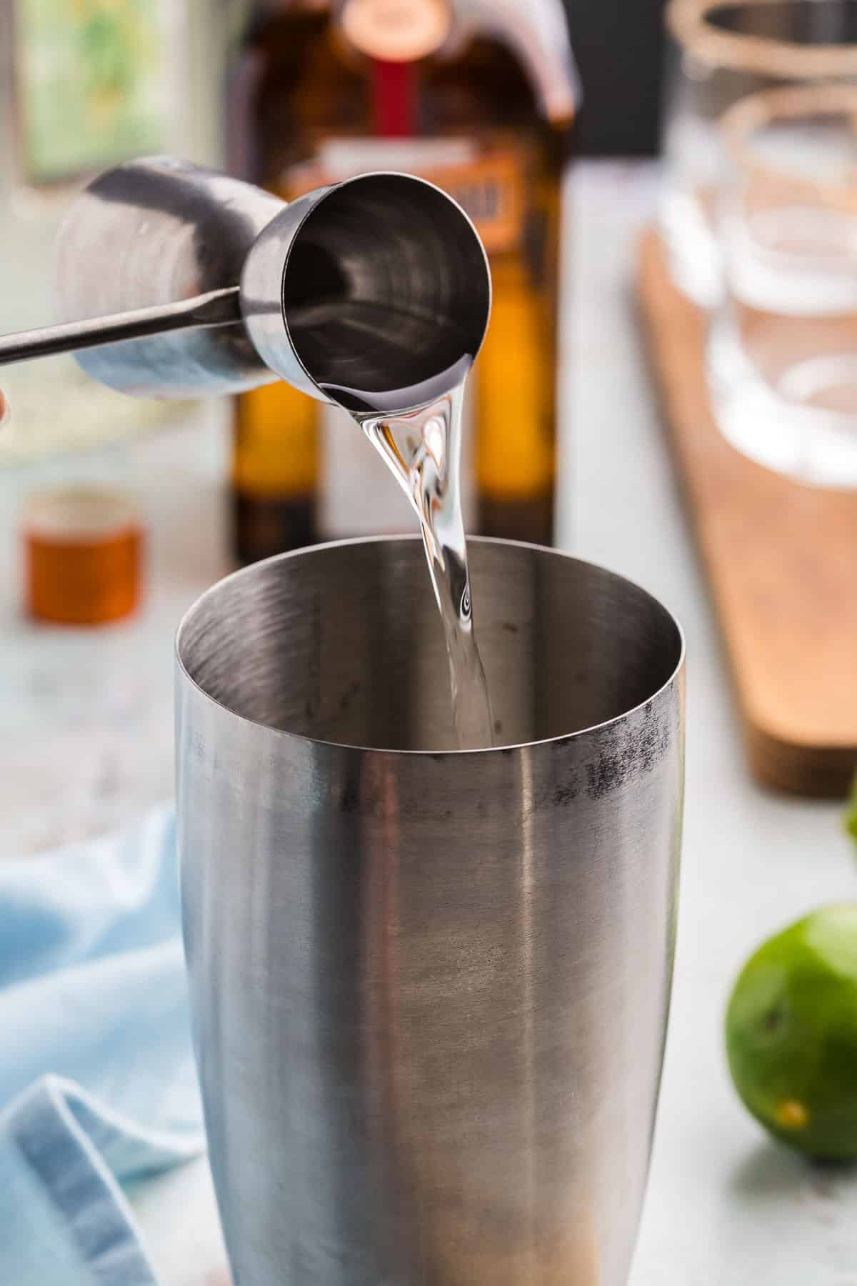 A side shot of a jigger of tequila being added to a stainless steel cocktail shaker.