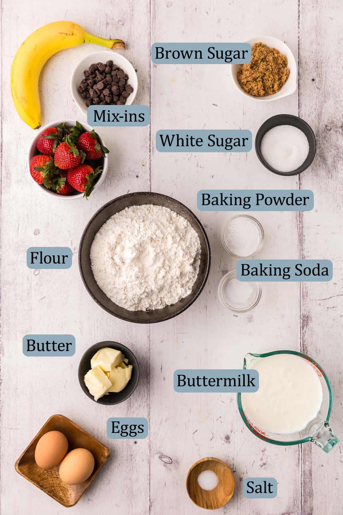 Overhead shot of the ingredients needed to make sheet pan pancakes. Ingredients include: flour, brown and white sugar, baking powder, baking soda, butter, buttermilk, eggs, and salt. Optional mix-ins like banana, strawberries, and chocolate chips.