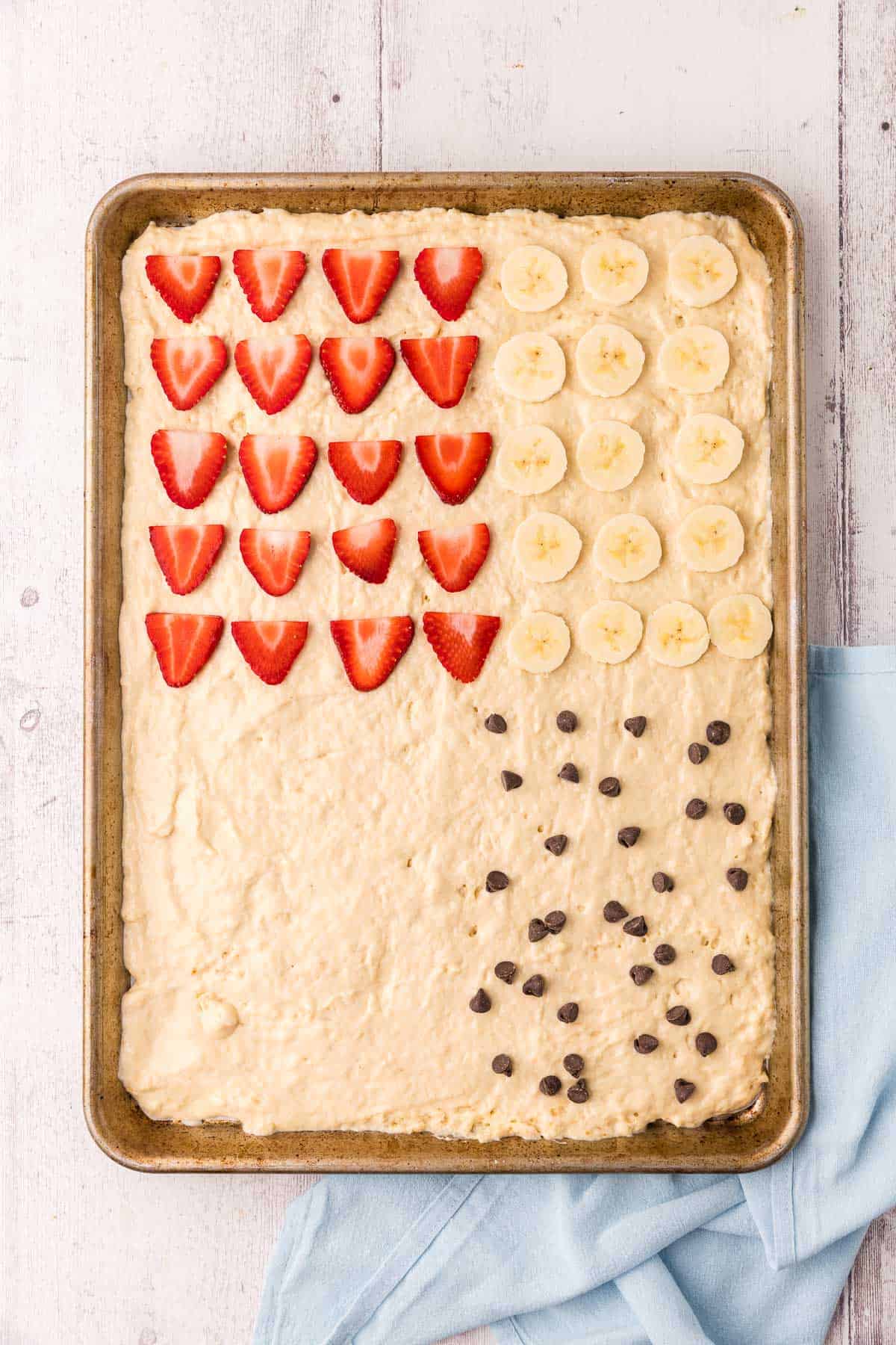overhead shot of the sheet pan with the pancake batter spread on. The top left corner is filled with cut strawberries. The top right corner is filled with cut banana, the bottom left is plain, and the bottom right has chocolate chips spread on it.