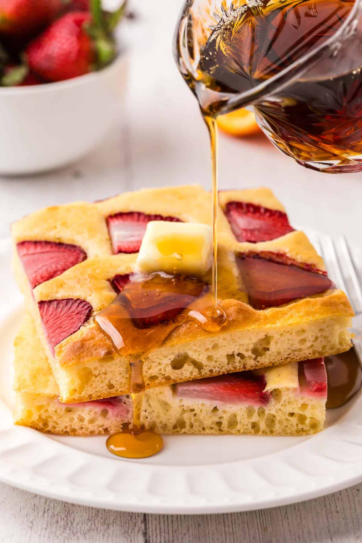 side shot of syrup being poured on a stack of sheet pan pancakes. The syrup is dripping down the front of the stack.