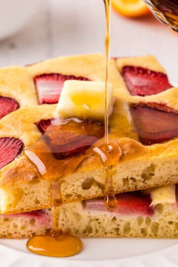 side shot of two strawberry sheet pan pancakes made in to a stack. A pat of butter is melting on the top and syrup is being poured on the stack.