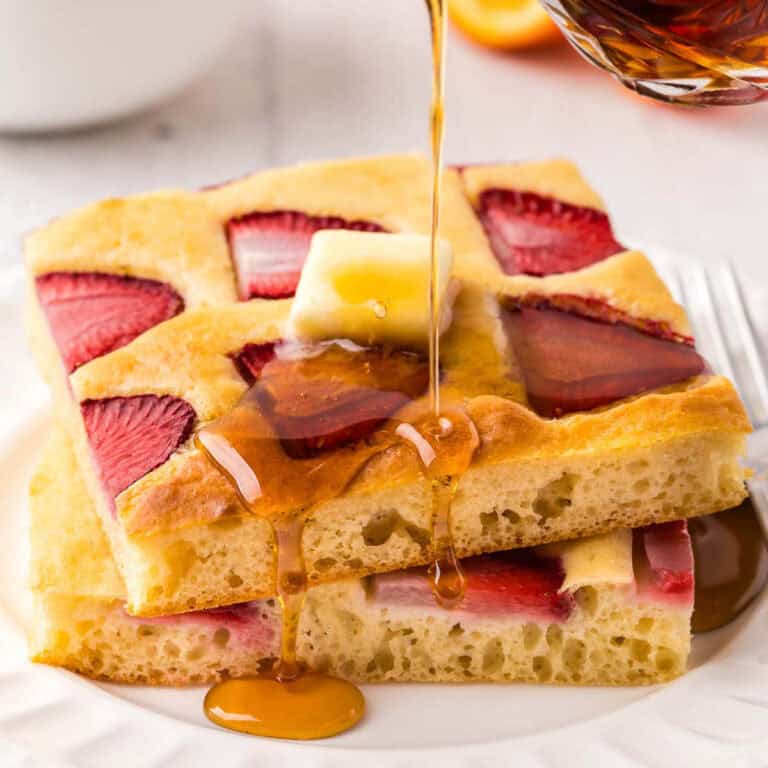side shot of two strawberry sheet pan pancakes made in to a stack. A pat of butter is melting on the top and syrup is being poured on the stack.