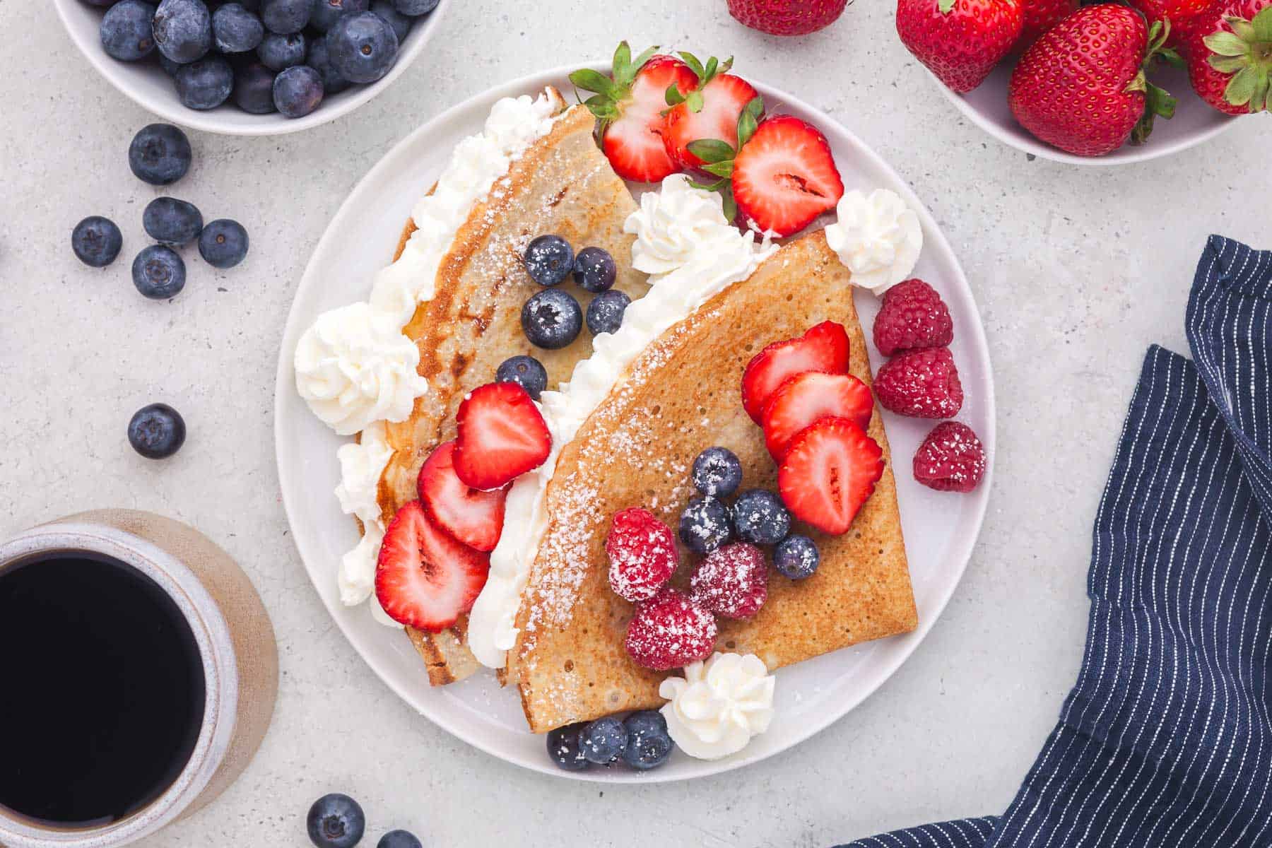 overhead shot of a plate of two crepes folded into triangles and filled with whipped cream. Fresh blueberries, strawberries, and raspberries sit on top along with a sprinkling of powdered sugar. Great for a brunch recipe.