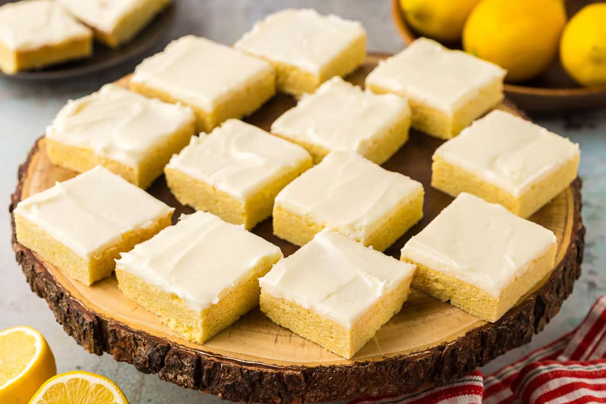 a high 45 degree angle shot of lemon cookie bars laid out on a wooden cake tray. Each cookie bar is iced with a lemon cream cheese frosting.